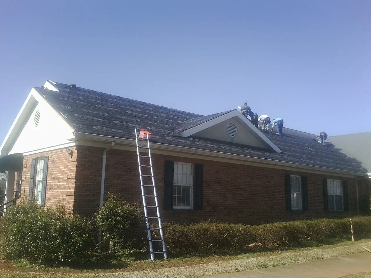 Crew tearing shingles off a brick ranch roof with a ladder set — Emergency Roof Repair work by our crew in Stilwell