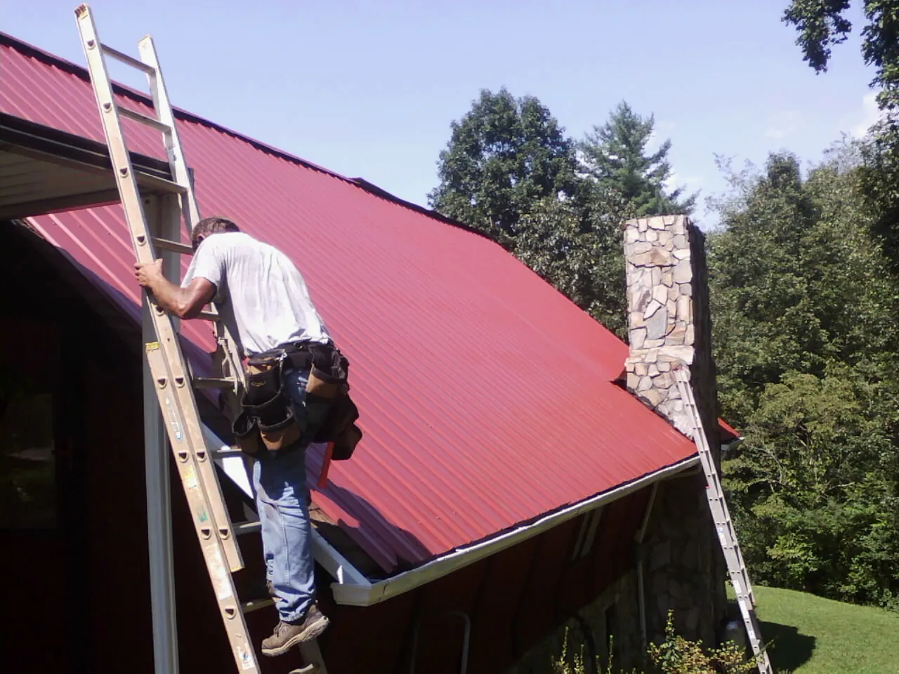 Roofer on a ladder beside a red metal roof and stone chimney — Soffit & Fascia Repair by Roofing Brothers in Stilwell